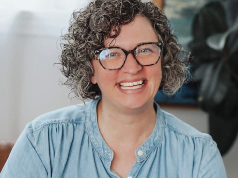 A professional image of Jennifer Ringgold, Director fo Faith Formation and Michigan Conference Mitten Project Program Director. Jenn is smiling and is wearing round framed glasses and a buttoned blue shirt.